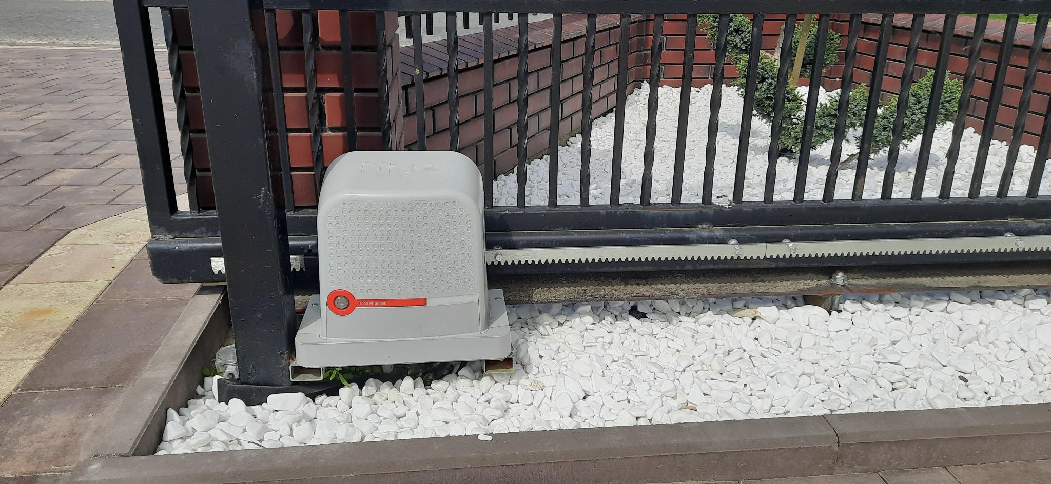 Szary automat do bramy przesuwnej Nice Hi-Speed, zamontowany przy czarnej, metalowej bramie. Białe kamienie dekoracyjne wokół. Widoczna listwa zębata.