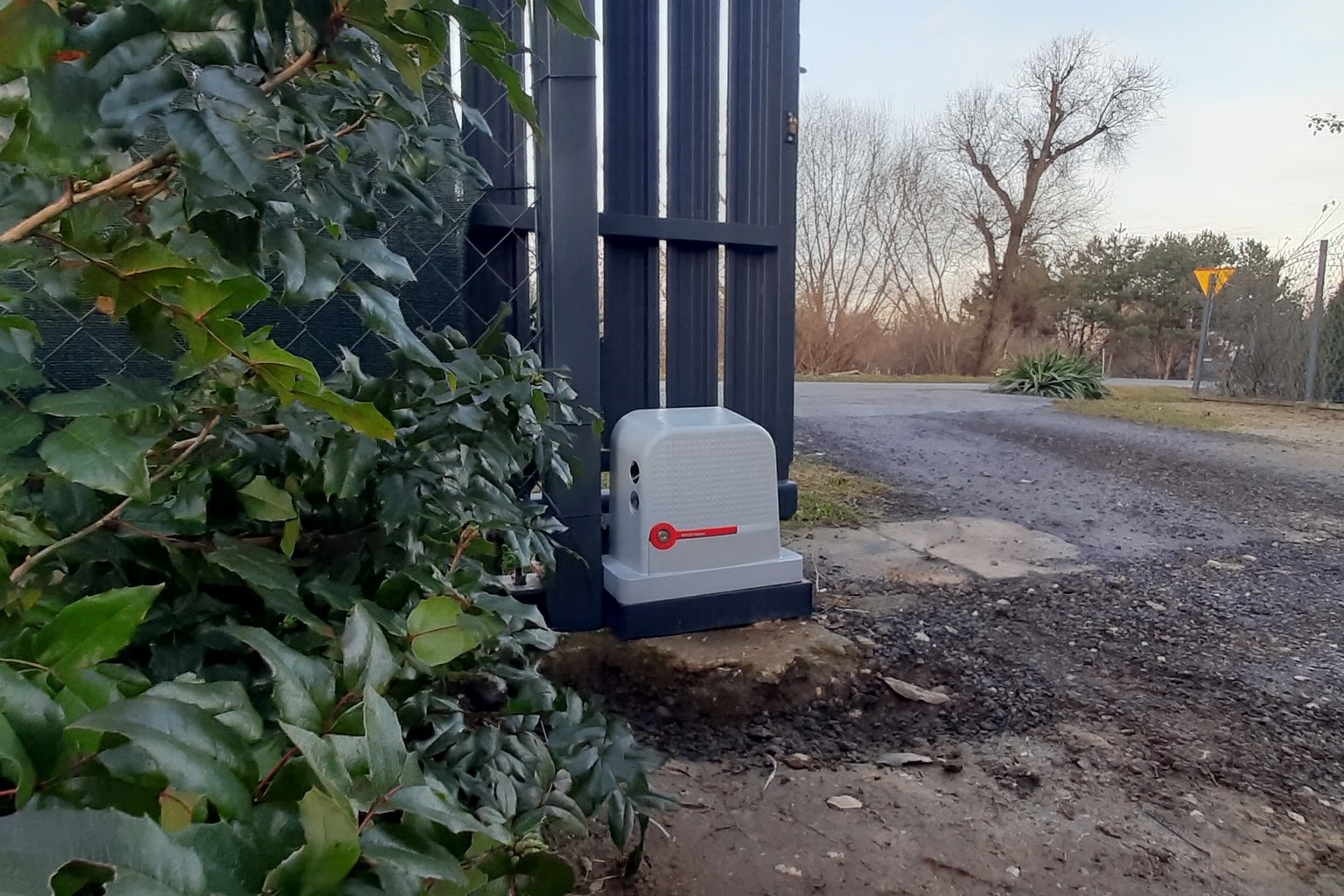 Szara automatyka bramy zamontowana przy metalowej bramie wjazdowej, obok bujnej zieleni. W tle droga i drzewa.