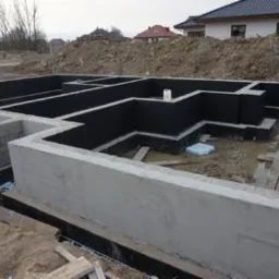 Surowe fundamenty domu jednorodzinnego z izolacją przeciwwilgociową, widoczne szalunki i teren budowy w tle. Detal: biały kanister wewnątrz fundamentów.