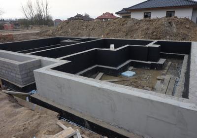 Surowe fundamenty domu jednorodzinnego z izolacją przeciwwilgociową, widoczne szalunki i teren budowy w tle. Detal: biały kanister wewnątrz fundamentów.