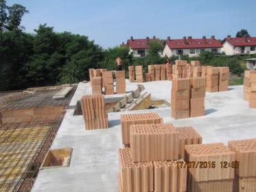 Wznoszenie ścian domu z cegły ceramicznej Porotherm na wylewce fundamentowej, widoczne zbrojenie i pustaki. W tle zabudowa jednorodzinna i zieleń.