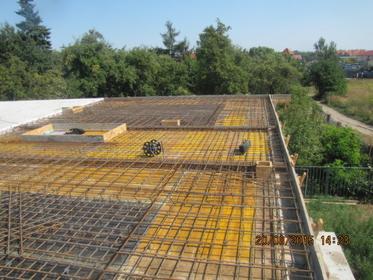 Zbrojenie stropu budynku w trakcie budowy, widoczne elementy szalunku i siatka zbrojeniowa, w tle zieleń drzew i budynki. Ujęcie z góry.