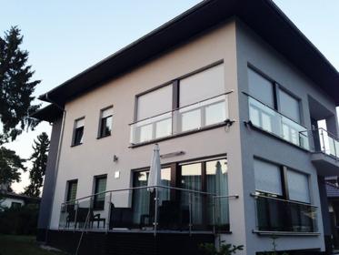 Dwukondygnacyjny dom z jasną elewacją, balkonami z balustradami ze szkła i metalu, roletami w oknach i tarasem z meblami ogrodowymi.