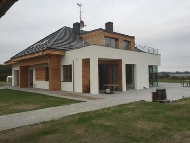 Nowoczesny dom z płaskim dachem i elementami drewna na elewacji, widok z ogrodu. Zwraca uwagę taras na piętrze z metalową balustradą i starannie zagospodarowany teren.