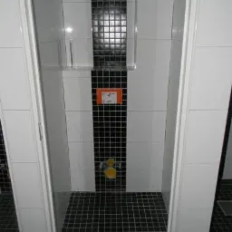 Wnętrze łazienki z czarno-białymi płytkami, mozaiką na ścianie i podłodze. Widoczny fragment kabiny prysznicowej oraz przycisk spłuczki.