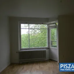 Pusty pok&oacute;j z parkietem, dużym oknem z widokiem na zieleń i grzejnikiem pod oknem. Ściany w jasnym odcieniu, gotowe do malowania.