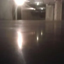 Błyszcząca, ciemna posadzka przemysłowa w hali magazynowej. Widoczne refleksy świetlne, betonowe filary w tle. Minimalistyczny, surowy wygląd.