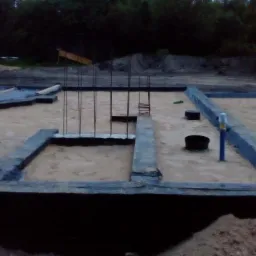 Fundamenty domu w trakcie budowy: betonowe ściany, stalowe pręty zbrojeniowe, piasek. Widok z góry, tło z zielenią.