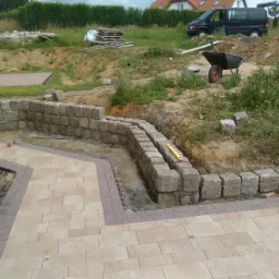 Budowa tarasu z kamienną ścianą oporową na nierównym terenie, widoczny wózek budowlany i palety, etap prac.