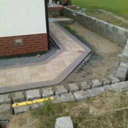 Fragment układanej ścieżki z jasnej kostki brukowej z szarym obrzeżem, przylegającej do ściany budynku z ceglaną podmurówką, obok wznoszony kamienny murek oporowy, poziomica na kamieniach.