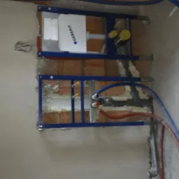 Montaż stelaża podtynkowego Geberit na ceglanej ścianie, widoczne rury instalacji wodnej w kolorze niebieskim i czerwonym. Ściana w trakcie prac wykończeniowych.