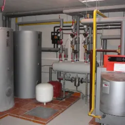 Kompleksowa kotłownia z dwoma dużymi zbiornikami, licznymi rurami, zaworami i manometrami. Czerwony piec grzewczy z elektronicznym panelem sterowania.