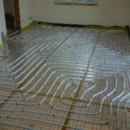 Białe rury ogrzewania podłogowego ułożone na folii z siatką w nowym budynku, widoczne okno i narzędzia. Etap instalacji przed wylewką.