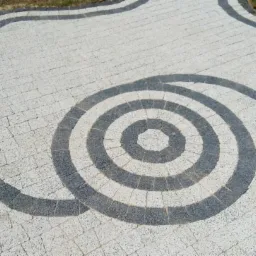 Ułożona ścieżka z kostki brukowej w odcieniach szarości i bieli, tworząca dekoracyjny wzór koncentrycznych okręgów i falistych linii.