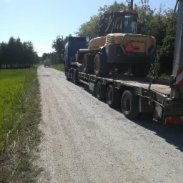 Żółta koparko-ładowarka Mecalac transportowana na niskopodwoziowej naczepie ciężarówki po szutrowej drodze w słoneczny dzień.