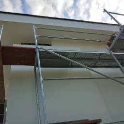 Elewacja budynku w trakcie prac wykończeniowych, widoczne rusztowanie i świeżo położony jasny tynk, błękitne niebo w tle.