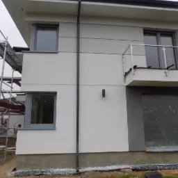 Elewacja dwukondygnacyjnego domu w trakcie prac wykończeniowych, z widocznymi rusztowaniami i zabezpieczonym folią otworem garażowym, prezentująca kombinację białego i szarego tynku.