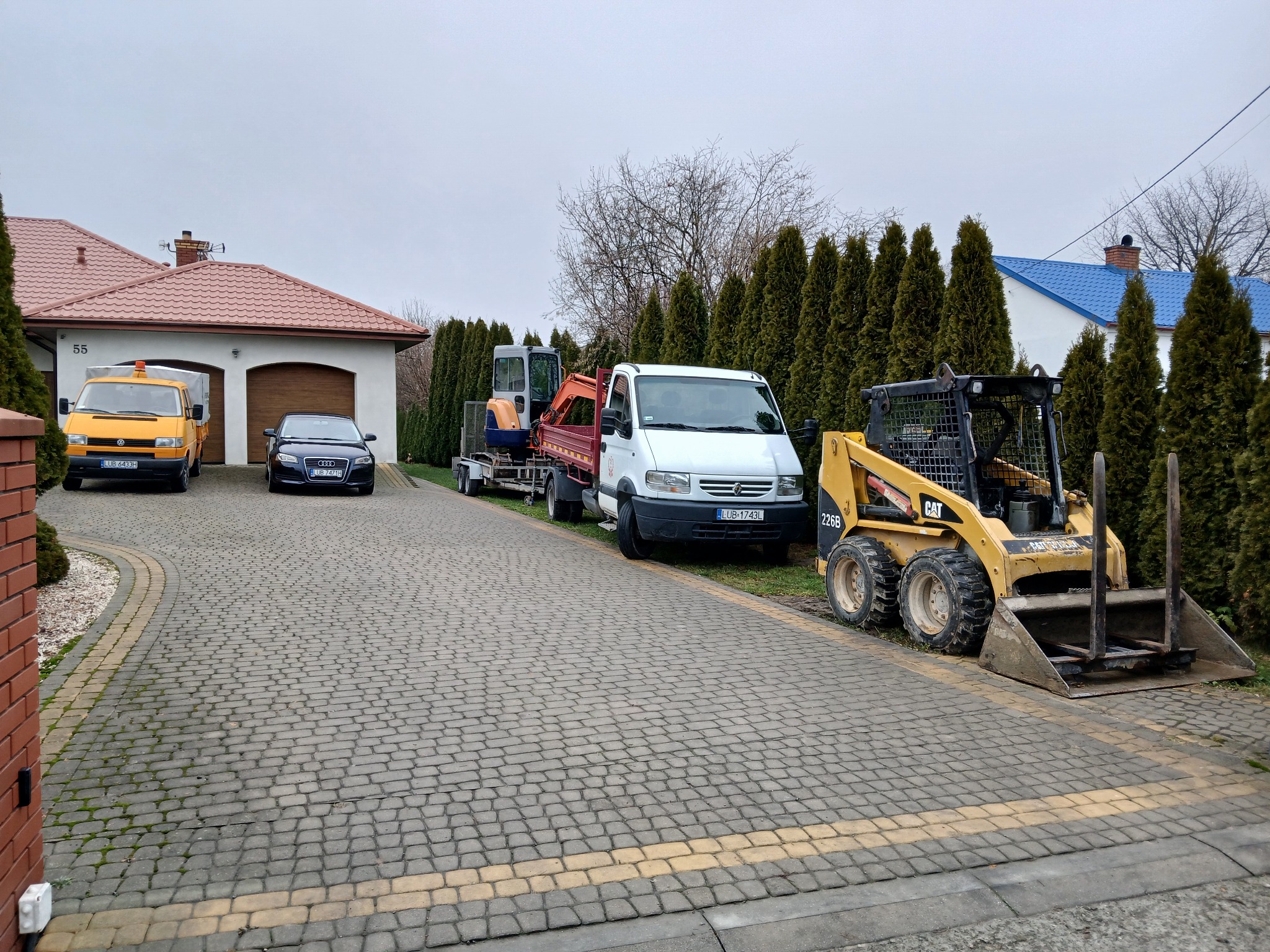 Żółty samochód dostawczy, czarny sedan, biały bus z przyczepą z mini koparką i żółta ładowarka CAT na posesji z kostki brukowej przed domem z czerwonym dachem i tujami w tle.