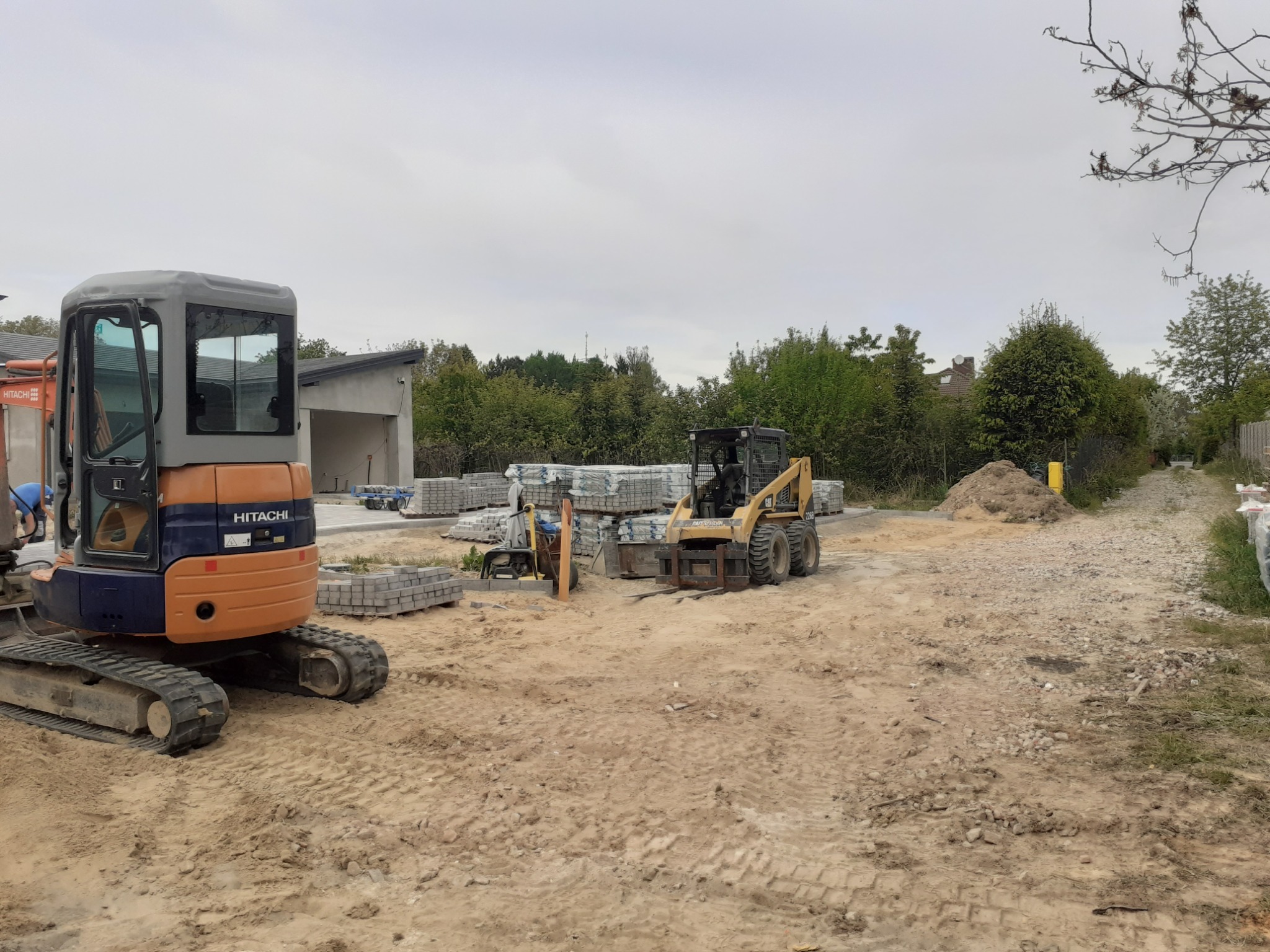 Pomarańczowy minikoparka Hitachi i żółta ładowarka Bobcat na placu budowy z przygotowanymi paletami kostki brukowej, widoczne ślady opon na piasku i częściowo ułożona kostka w tle.