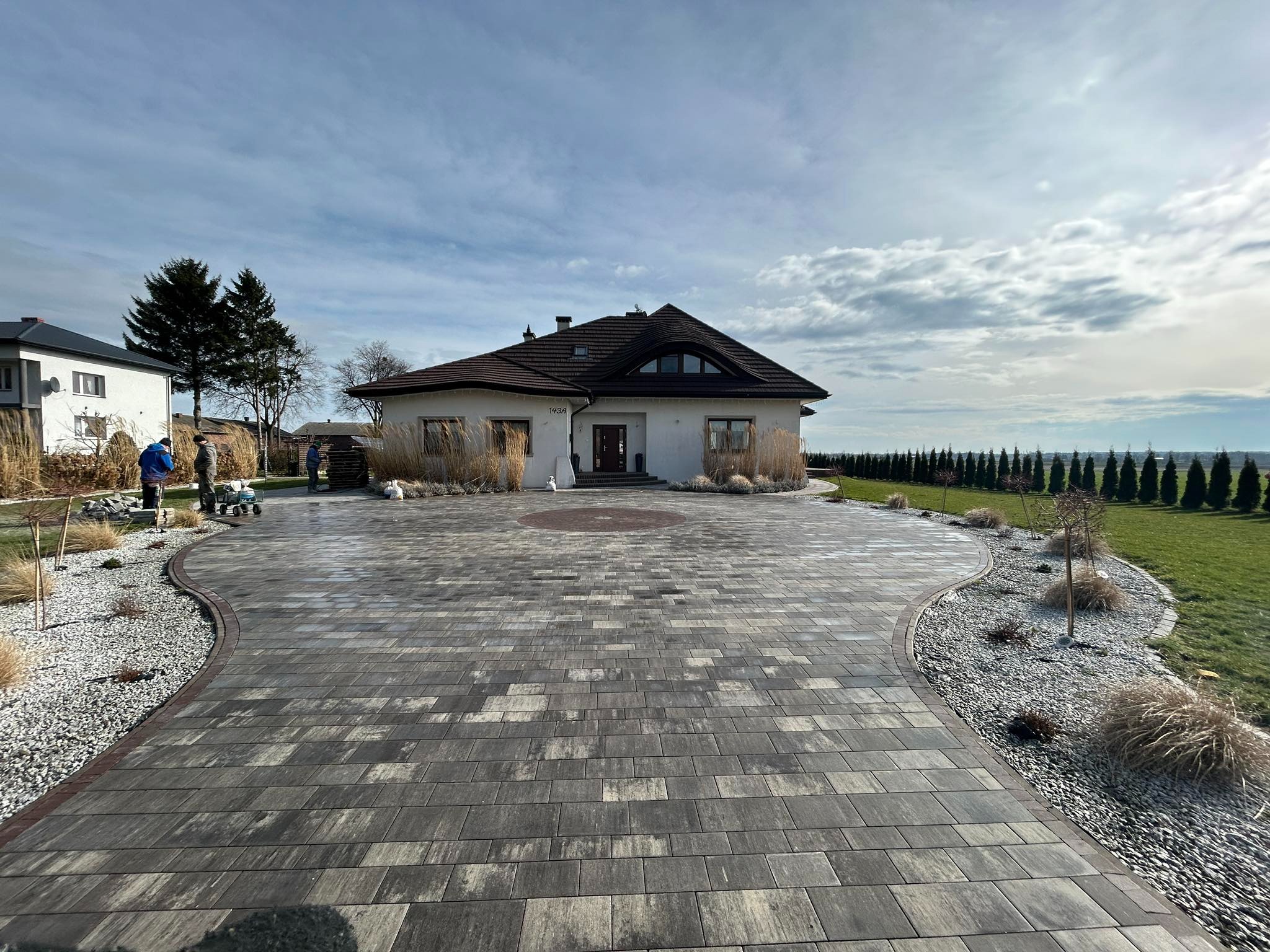 Szeroki, zakrzywiony podjazd z szarej kostki brukowej prowadzący do nowoczesnego domu z brązowym dachem, otoczony ozdobnymi trawami i białym żwirem, widoczni pracownicy w oddali.