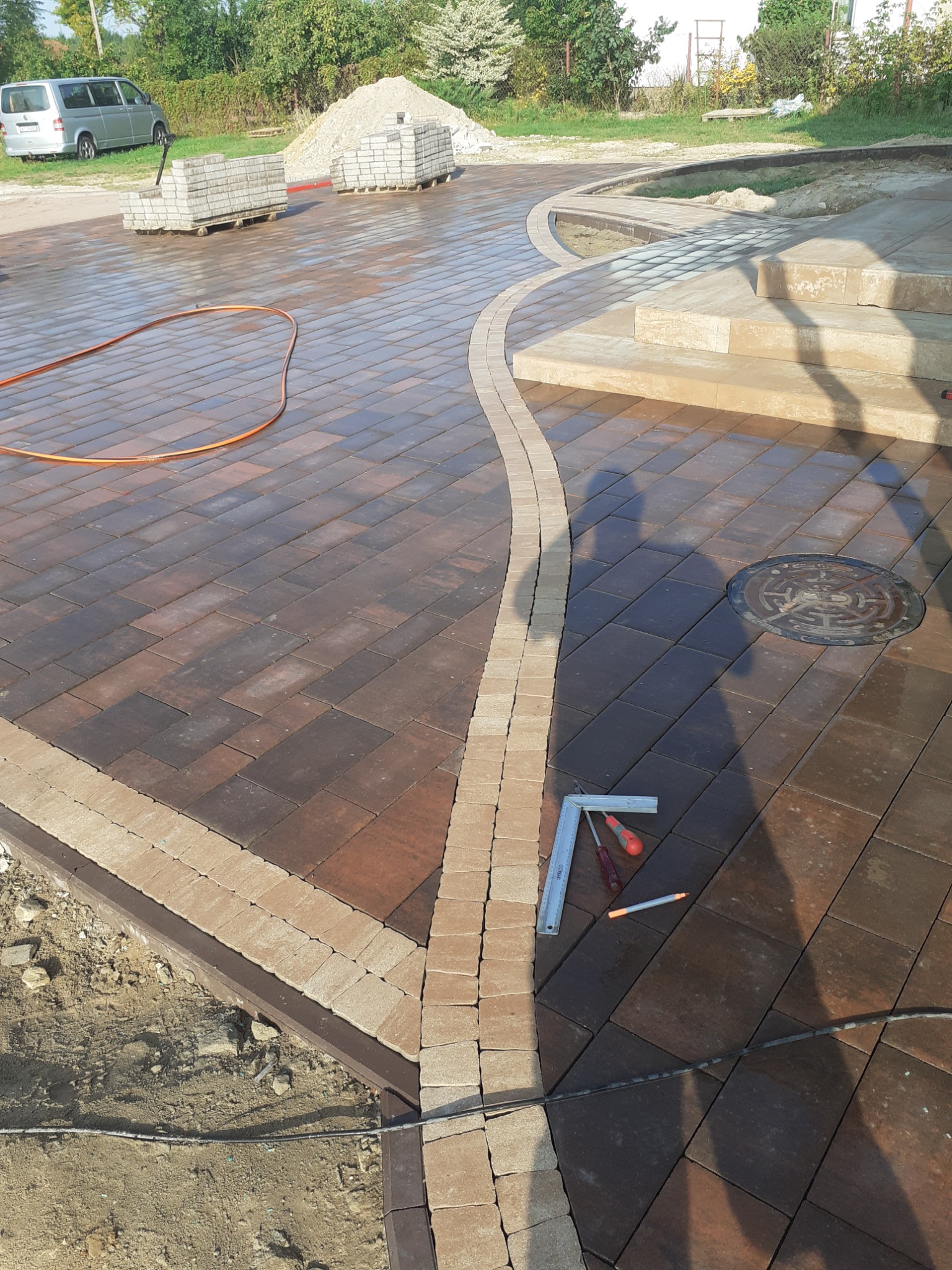 Ułożona kostka brukowa w odcieniach brązu z jasnym pasem dekoracyjnym, widoczne narzędzia brukarskie, studzienka kanalizacyjna i schody wejściowe do domu.