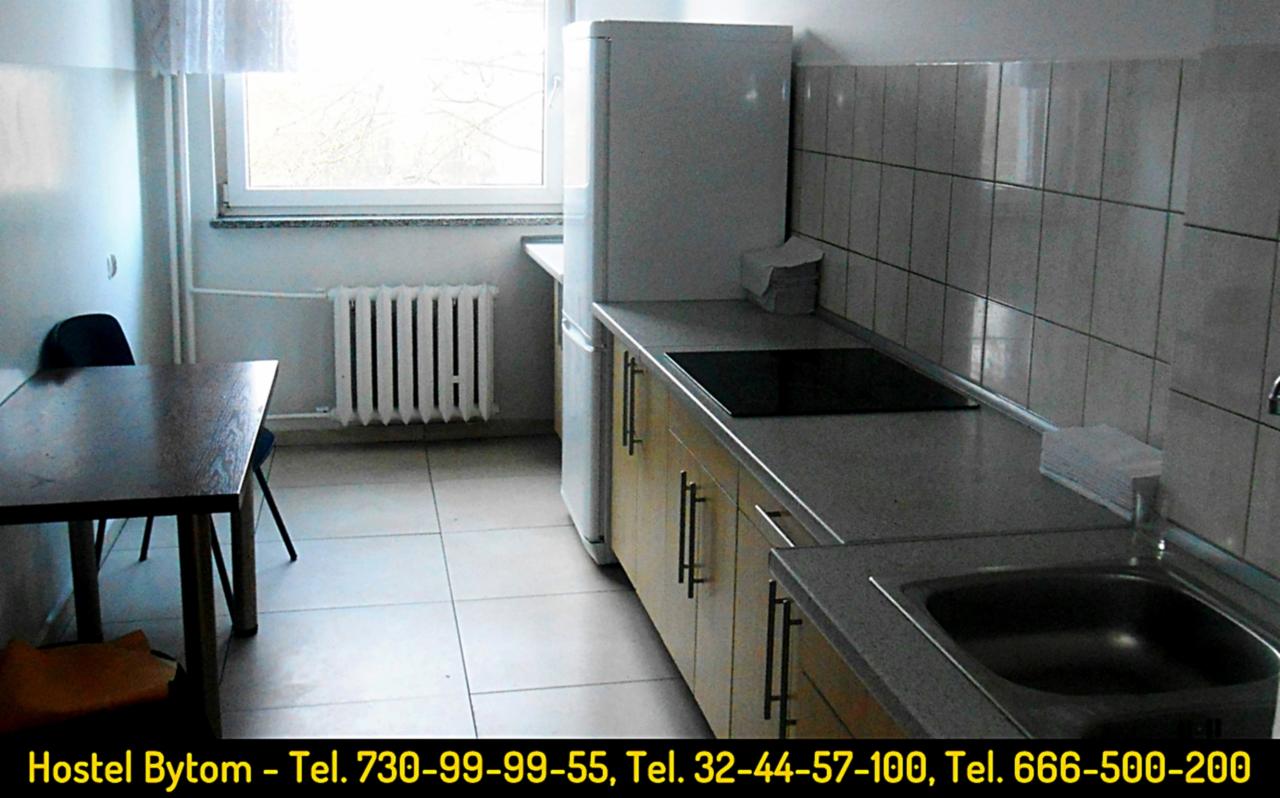 Wnętrze kuchni w hostelu: stół z krzesłami, grzejnik pod oknem, zabudowa kuchenna z płytą grzewczą i zlewem, biała lodówka.