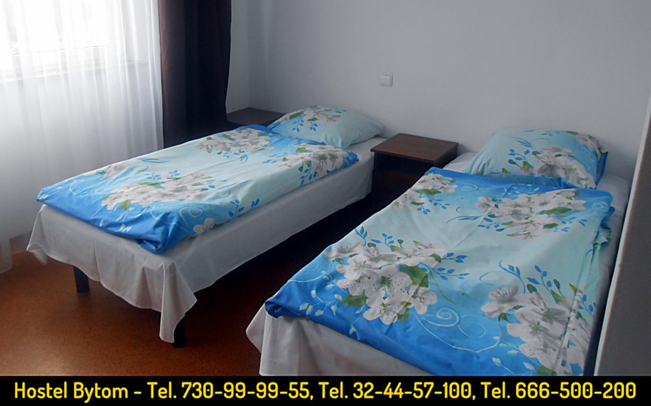 Pokój w hostelu z dwoma pojedynczymi łóżkami, pościel w kwiatowy wzór, stolik nocny między łóżkami, widok na okno z zasłonami.