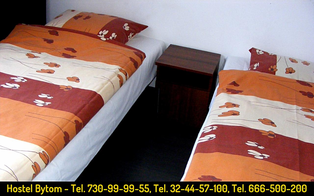 Pokój w hostelu z dwoma łóżkami pojedynczymi, nakryte pościelą w paski w odcieniach brązu, pomarańczy i bieli z motywem kwiatowym, pomiędzy nimi ciemnobrązowa szafka nocna, czarna podłoga.