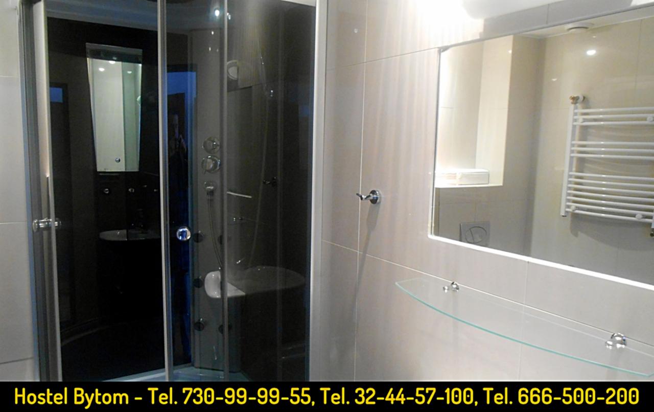 Łazienka w hostelu: kabina prysznicowa z hydromasażem, lustro, szklana półka i grzejnik drabinkowy na ścianie z jasnych płytek.