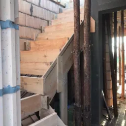 Wnętrze w trakcie budowy z drewnianymi schodami tymczasowymi, ceglaną ścianą i drewnianymi podporami, wpadające promienie słoneczne.