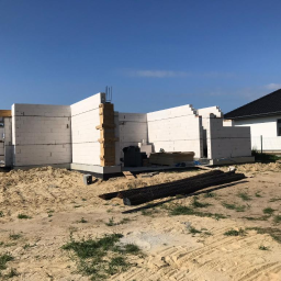 MAGBUD Bartłomiej Kardziejonek - Wznoszenie ścian parteru z białych bloczków na fundamencie, widoczne zbrojenia i deski szalunkowe, w tle dom jednorodzinny z ciemnym dachem.