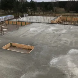 Zalany betonowy fundament z drewnianym szalunkiem na obrzeżach i otworem, widok z góry na plac budowy w pochmurny dzień.