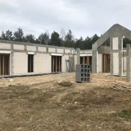 Surowy stan domu jednorodzinnego z betonu komórkowego w trakcie budowy, z widocznymi drewnianymi stemplami w otworach okiennych, bloczkami betonowymi na pierwszym planie i lasem w tle.