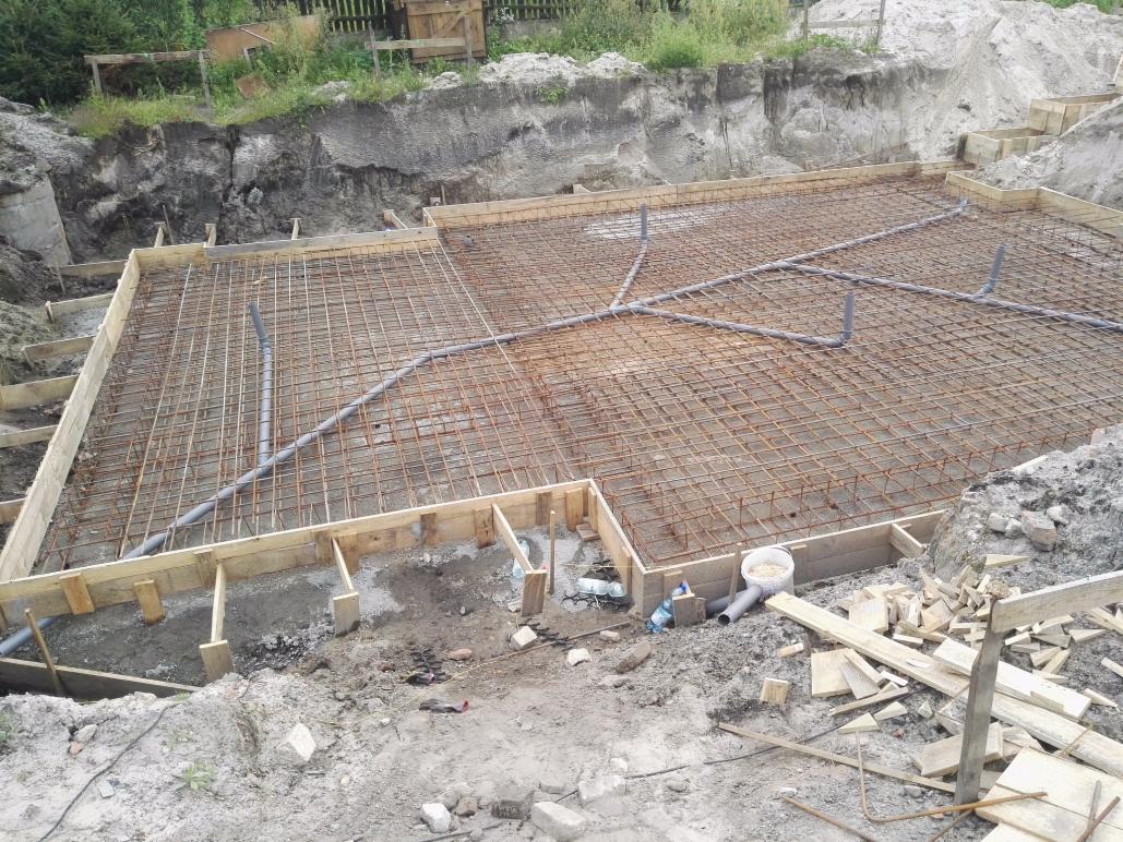 Zbrojenie fundamentów domu jednorodzinnego z widocznymi rurami instalacji wodnej, szalunek z desek i wykop ziemny w tle.