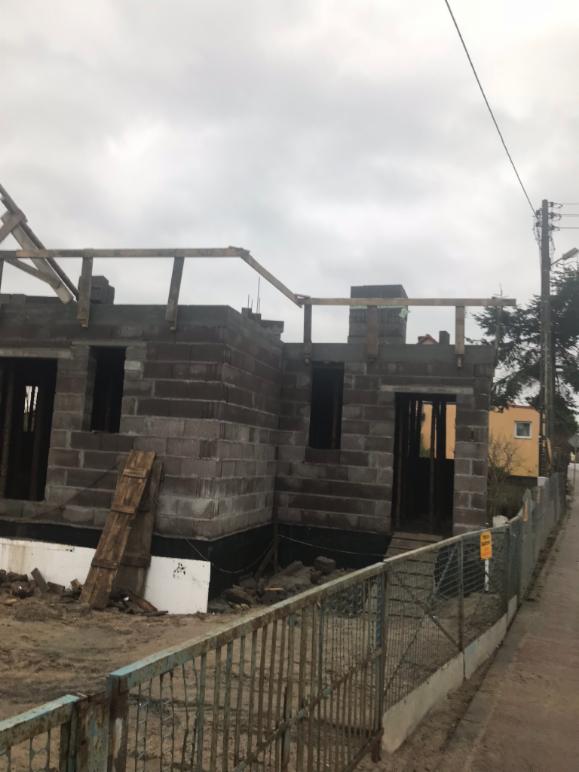 Surowy stan budynku z szarej cegły z drewnianą konstrukcją dachu w trakcie budowy, widoczne okna i drzwi bez ram, ogrodzenie w tle, pochmurne niebo.