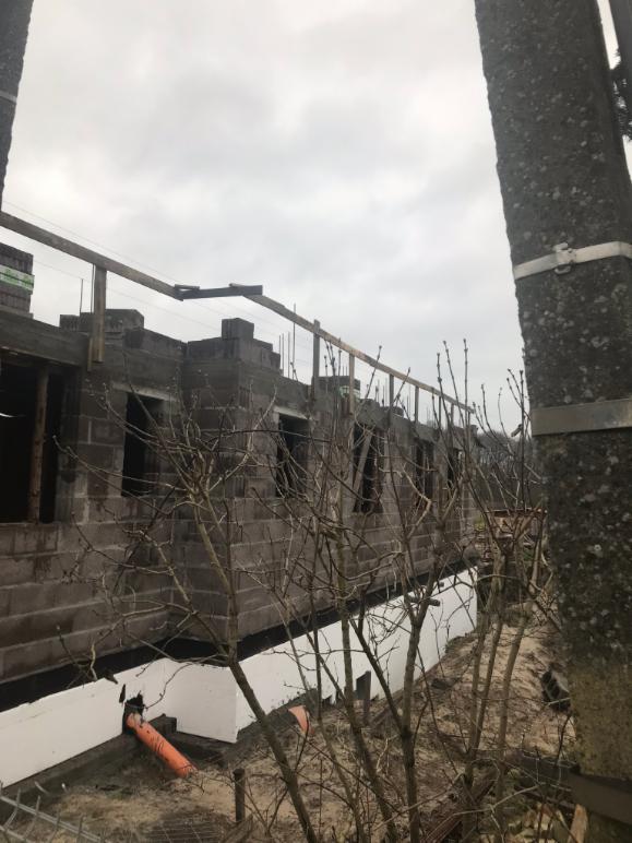 Surowy stan budynku z bloczków, widoczne okna bez ram, zabezpieczenia na dachu, izolacja fundamentów i rury odprowadzające wodę, pochmurne niebo w tle.