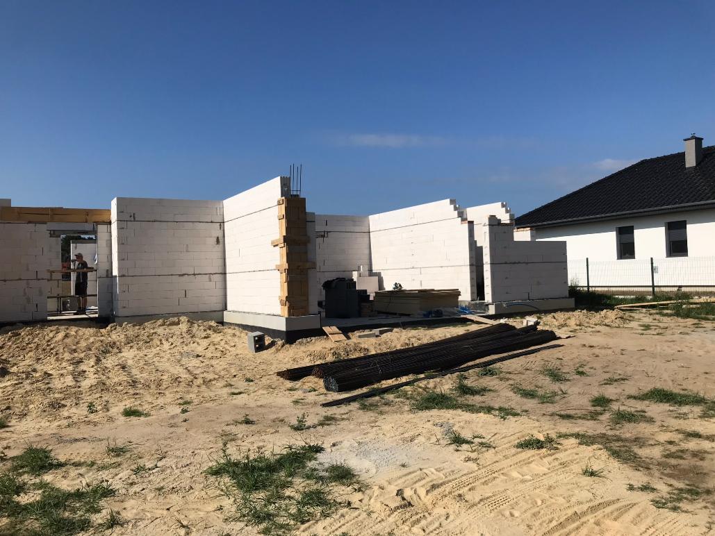 Wznoszenie ścian parteru z białych bloczków na fundamencie, widoczne zbrojenia i deski szalunkowe, w tle dom jednorodzinny z ciemnym dachem.