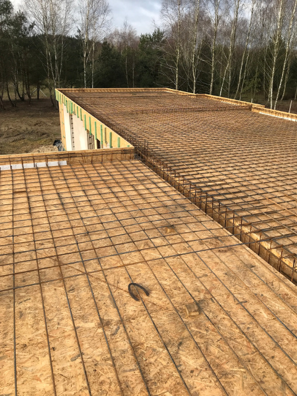 Wiązanie zbrojenia na deskowaniu stropu budynku w trakcie budowy, widoczny fragment ściany z pionowymi elementami drewnianymi, w tle las.