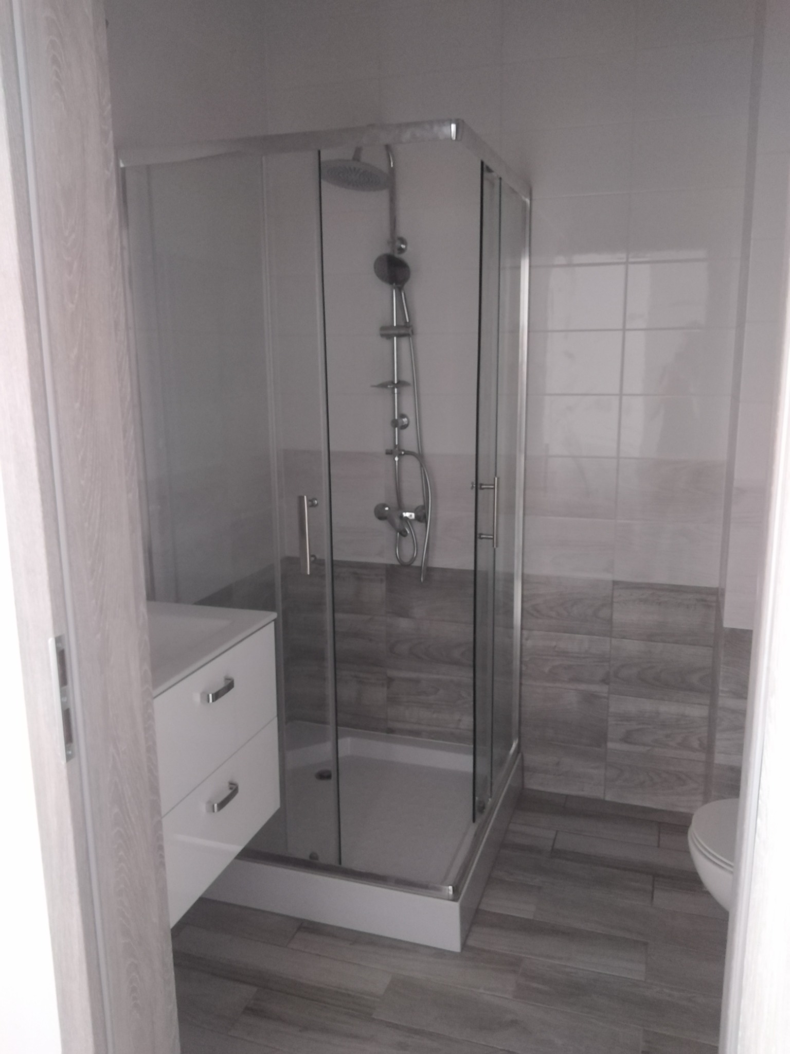 Kabina prysznicowa z deszczownicą i panelem natryskowym, umywalka z szafką, szaro-białe płytki imitujące drewno na ścianie i podłodze, widok zza otwartych drzwi.