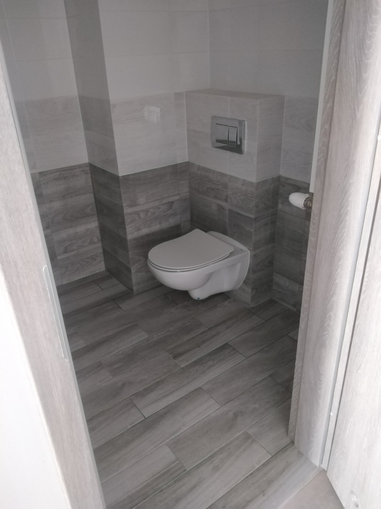 Nowoczesna łazienka z podwieszaną toaletą, wykończona płytkami imitującymi drewno w odcieniach szarości i bieli, widok zza uchylonych drzwi.