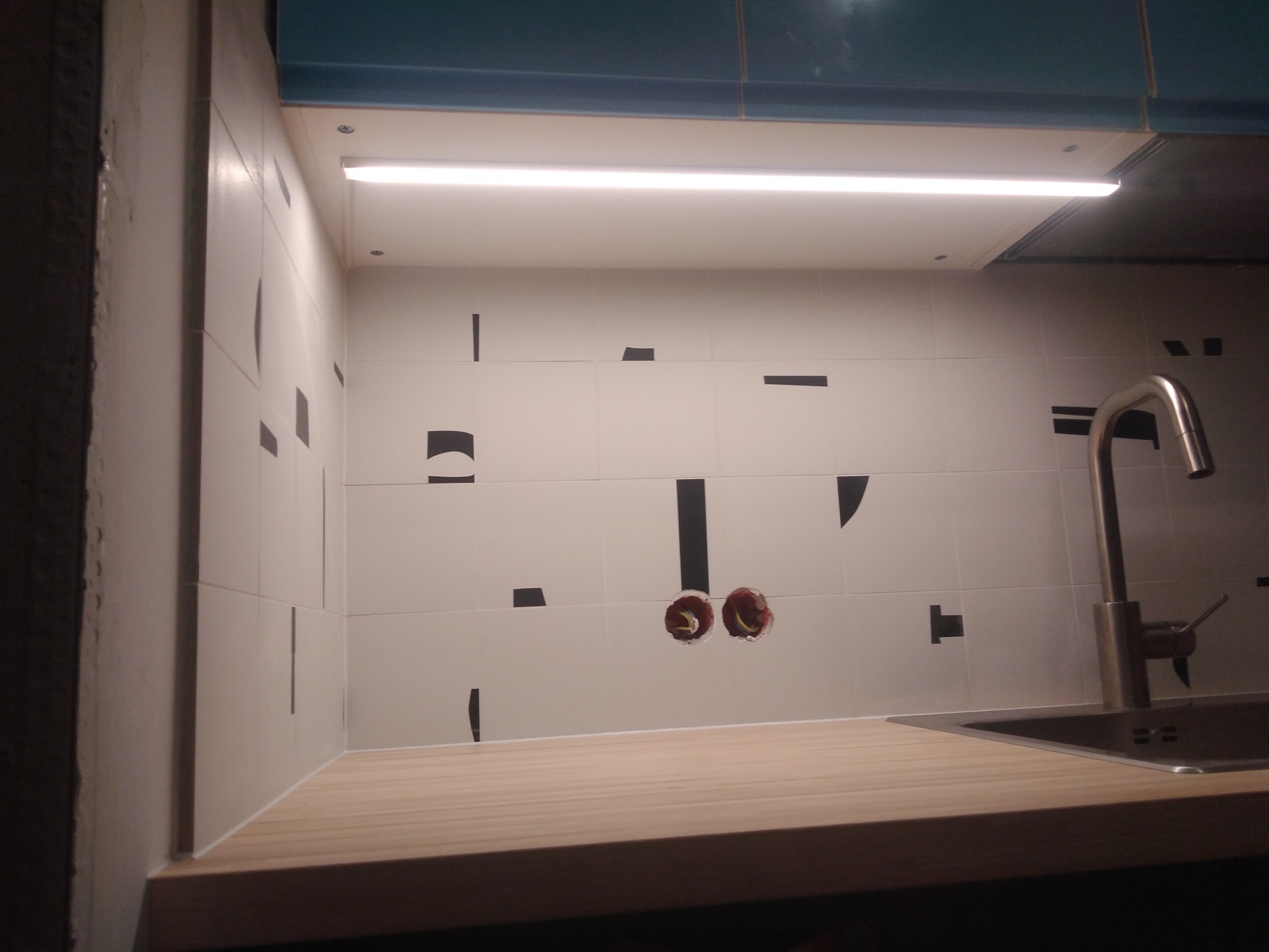 Białe płytki z czarnymi, geometrycznymi wzorami na ścianie nad blatem kuchennym, widoczne otwory na instalację elektryczną, bateria zlewozmywakowa i oświetlenie LED pod szafkami.