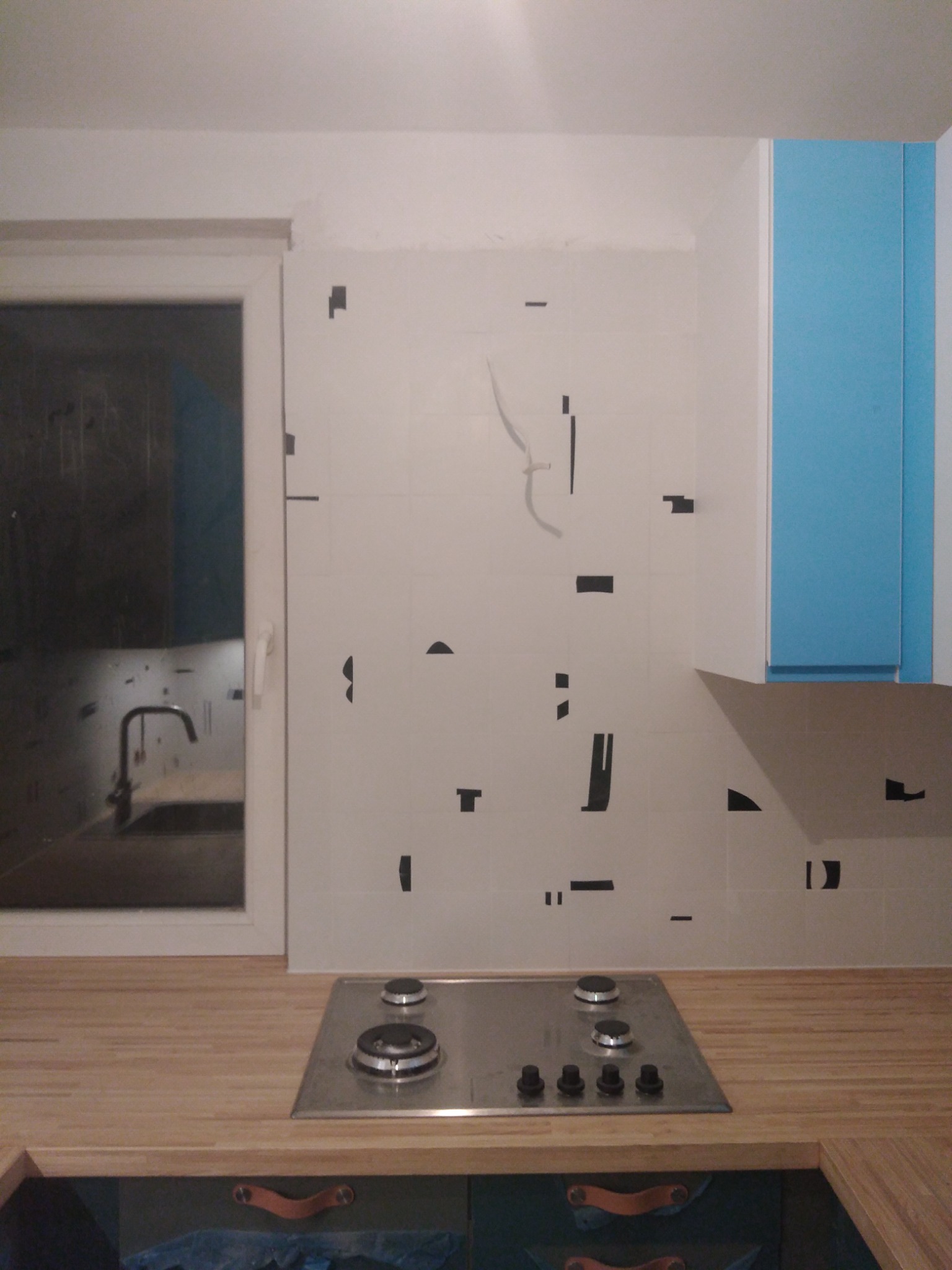 Fragment ściany w kuchni z częściowo położonymi białymi płytkami, zabezpieczonymi czarną taśmą, widoczna kuchenka gazowa na drewnianym blacie i okno.