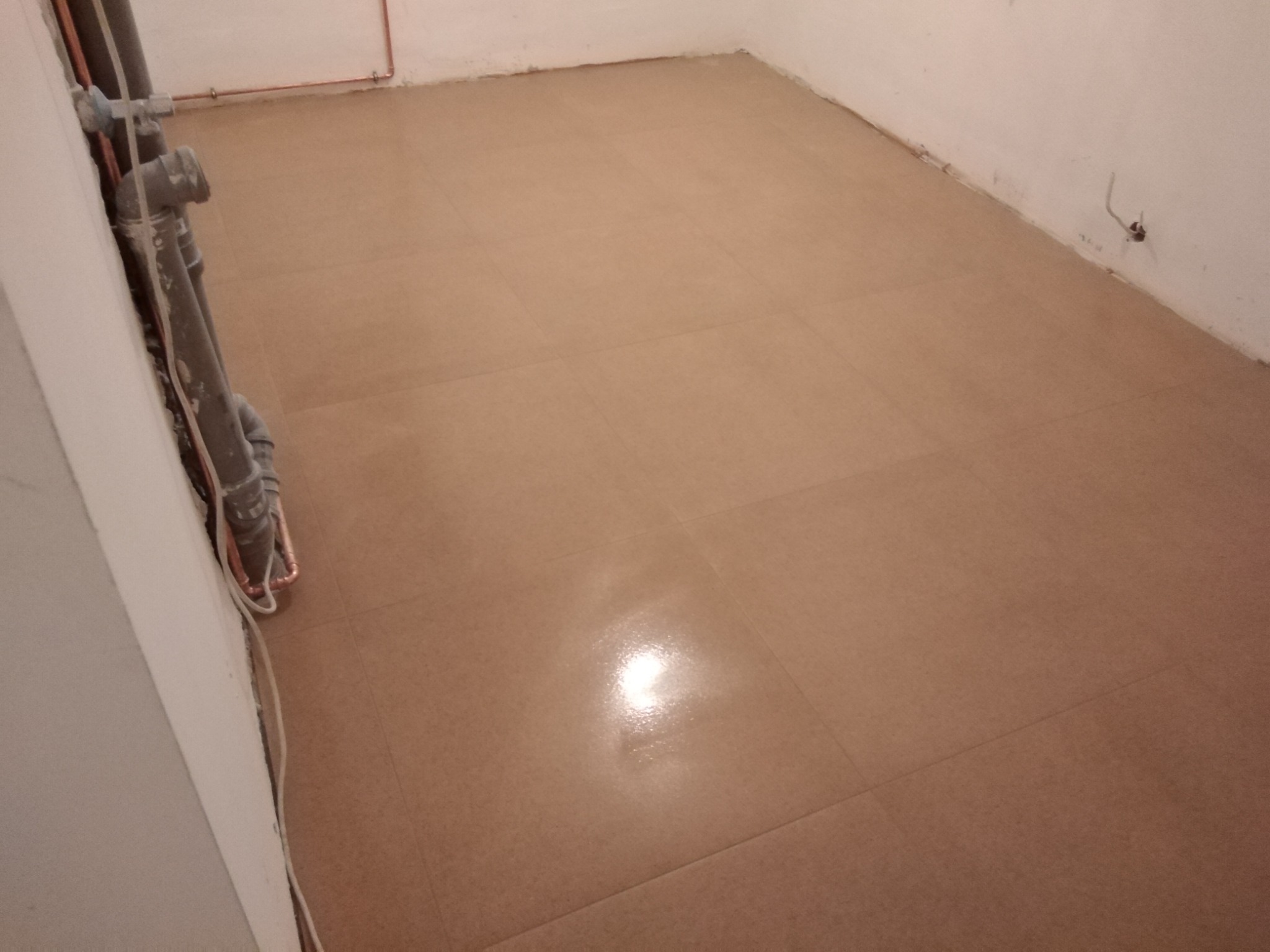 Ułożona, beżowa podłoga z płytek w pomieszczeniu w trakcie remontu, widoczne rury przy ścianie.