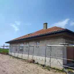 Dom w trakcie budowy z częściowo ułożonym pokryciem dachowym z dachówki ceramicznej w kolorze ceglastym, widoczne rusztowania i niezabezpieczone okna.
