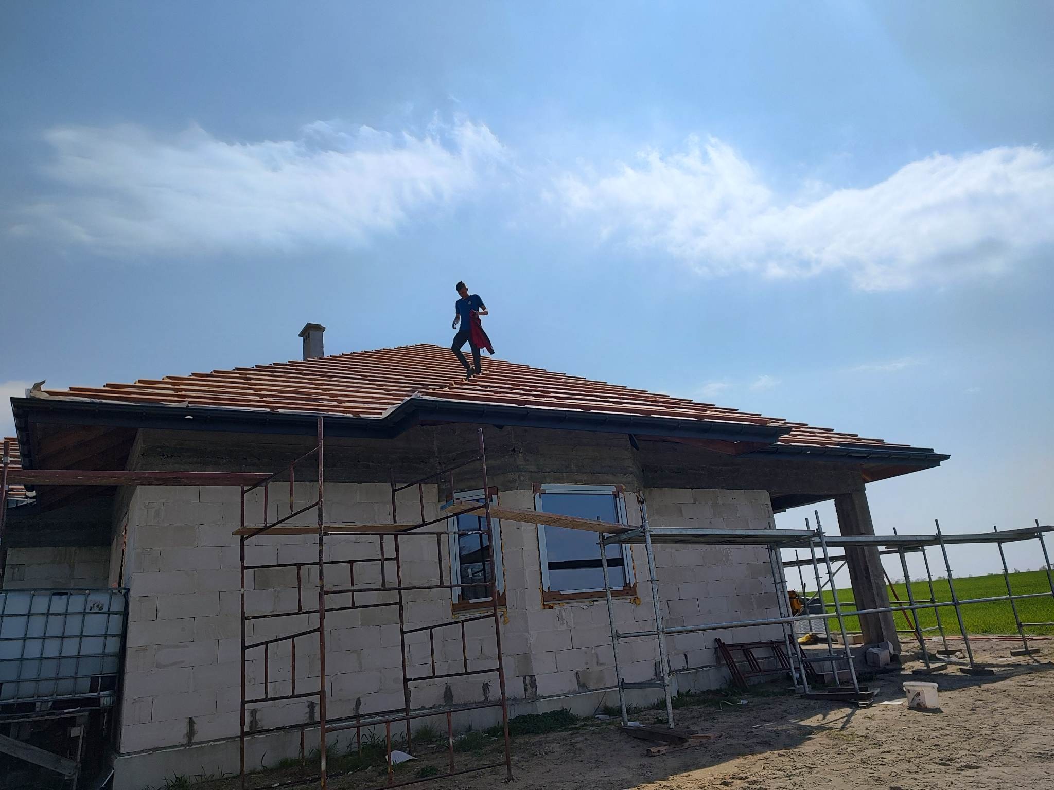 Dekarz na dachu domu jednorodzinnego w trakcie krycia czerwoną dachówką, widoczne rusztowania i ściany z betonu komórkowego.