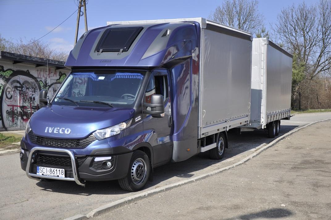 Niebieski Iveco z naczepą na parkingu, widoczne detale: relingi, wysoka kabina z oknem dachowym. W tle budynek z graffiti i drzewa.