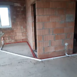 Surowy stan budynku z widoczną instalacją ogrzewania podłogowego w postaci białych rur i czerwonych przewodów, ułożonych na szarym, betonowym podłożu przy ścianach z cegły dziurawki.