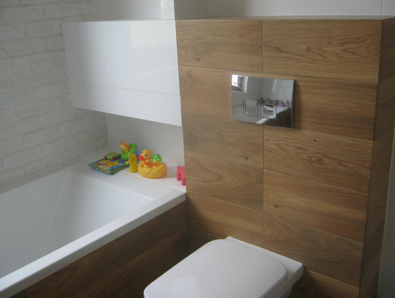 Nowoczesna łazienka z drewnopodobnymi płytkami na ścianie, wisząca toaleta z chromowanym panelem spłukującym, biała wanna obudowana drewnem, biała szafka wisząca i zabawki dla dzieci na wannie.