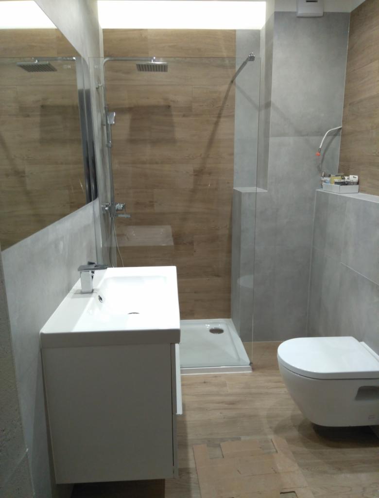 Nowoczesna łazienka z kabiną prysznicową typu walk-in, umywalką z szafką w kolorze białym, toaletą podwieszaną i drewnopodobnymi płytkami na podłodze i ścianie w strefie prysznica.