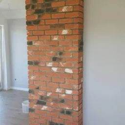 Dekoracyjny filar z czerwonej cegły z czarnymi i białymi akcentami, na tle szarej ściany i drewnianej podłogi.